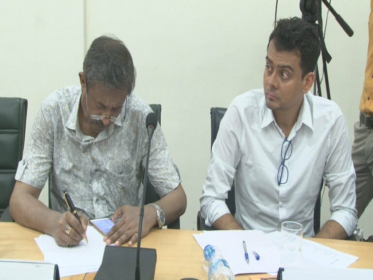 Two participants at a seminar table, the senior man writing notes while a younger colleague in a white shirt listens.