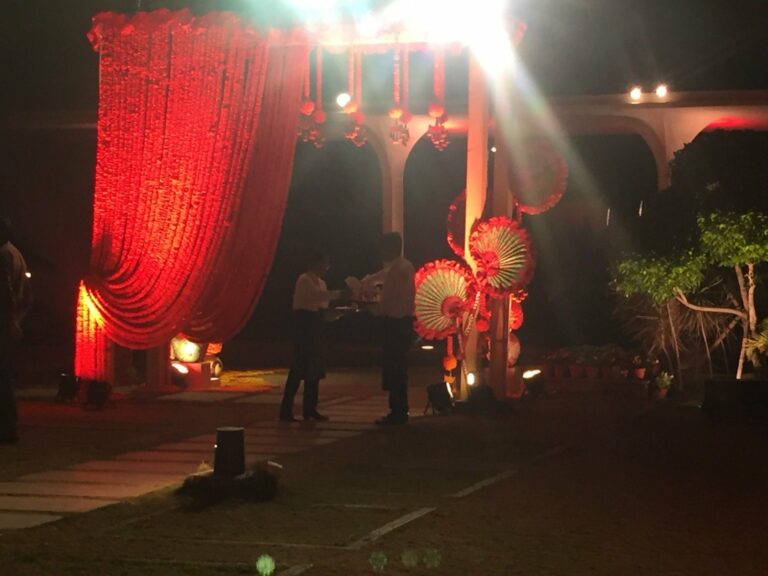 Evening view of the event venue: a red draped entrance lit with lamps and decorative parasols, guests standing outside on the lawn.