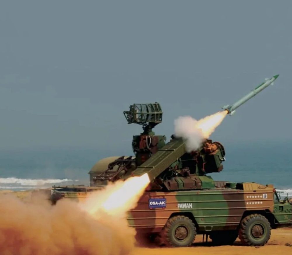 A camouflaged six-wheeled OSA-AK air-defence vehicle marked PAWAN launches a surface-to-air missile from a beach, exhaust flame and dust streaming behind it.