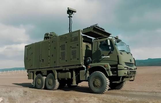 A dark green six-wheeled military truck carrying a boxed equipment shelter, with a mast-mounted radar and electro-optical sensor head raised above the cab.