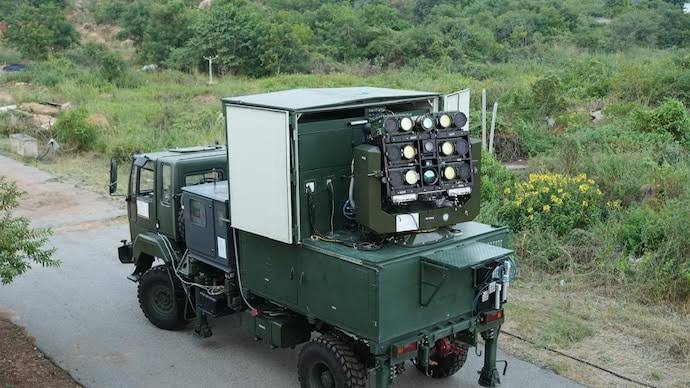 A truck-mounted laser directed-energy weapon on a hillside track, its cluster of nine circular beam apertures aimed skyward from an open equipment bay.