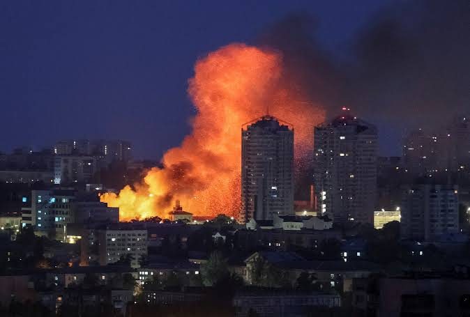 A wall of orange fire and black smoke rises between residential tower blocks on the Kyiv skyline at night, lighting the streets below.