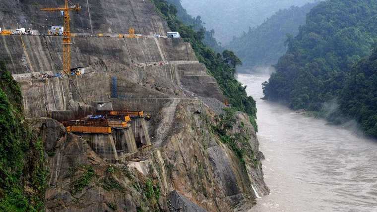 Construction cut into a steep gorge wall beside a fast river: concrete faces, tower cranes and excavators.